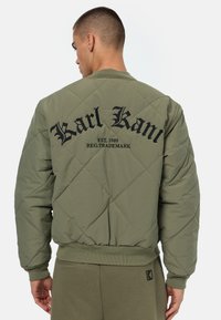 Vatteret olivengrøn bomberjakke med ribbede manchetter og talje. Har sort broderet logo og tekst på ryggen.