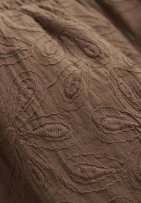 Brązowa tkanina haftowana z kwiatowymi wzorami o wypukłych, teksturowanych detalach. Gładkie tło uzupełnia skomplikowane wzory.