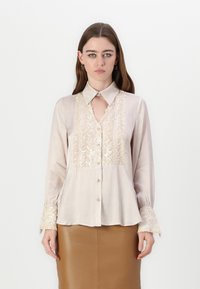 Blusa di seta color beige chiaro, con dettagli ricamati in oro, scollatura a chiave e maniche lunghe con polsini testurizzati.