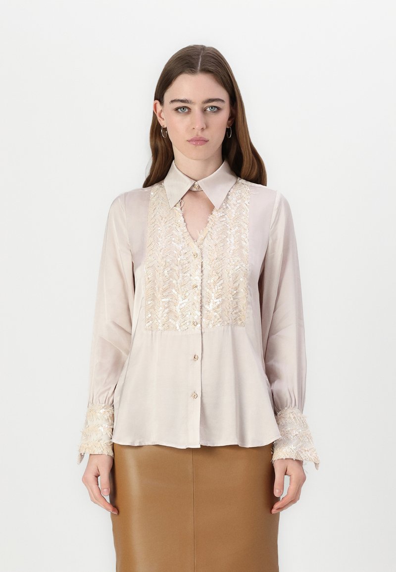 Blusa di seta color beige chiaro, con dettagli ricamati in oro, scollatura a chiave e maniche lunghe con polsini testurizzati.