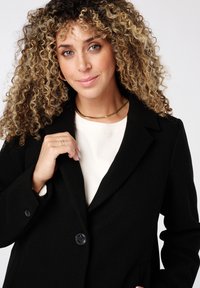 Manteau noir en mélange de laine avec un col cranté, fermeture à un bouton et deux poches latérales. Le modèle porte un sweat-shirt crème en dessous.