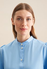 Helles blaues Bluse mit hohem Kragen, Knopfleiste mit drei Knöpfen und Puffärmeln. Glattes Stoffmaterial mit subtiler glänzender Oberfläche und taillierter Passform.