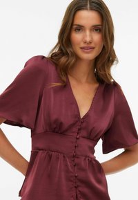 Vinrød satengbluse med V-hals, korte flutter-ermene og knappelukking foran. Glatt tekstur med innsvingt midje og utflared bunn.