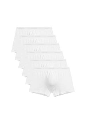 Muskułowaty mężczyzna o jasnej karnacji i blond włosach, ubrany w białe bokserki, stojący na tle jednolitego jasnego tła.