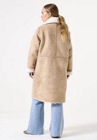 Manteau en shearling beige avec un col en fausse fourrure blanche, coupe ample et manches longues ; associé à un jean évasé bleu clair. Texture lisse, coutures minimales.