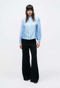Svetlomodrá blúzka s dlhým rukávom, vysokým golierom a zapínaním na gombíky; v kombinácii s čiernymi širokými nohavicami; hladká látka s jemnou textúrou.