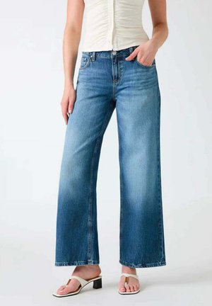 Donna che indossa jeans blu sbiadito a gamba larga, top bianco senza maniche arricciato e sandali bianchi con tacco e anello per le dita, in piedi davanti a uno sfondo neutro.