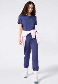 Mujer con camiseta y pantalones de Champion en color navy, sudadera rosa atada a la cintura, caminando con zapatillas blancas y rosas frente a un fondo neutro.