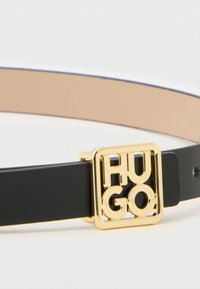 Czarny skórzany pasek z kwadratową, złotą klamrą z wytłoczonymi literami "HUGO". Gładka faktura i minimalistyczny design.