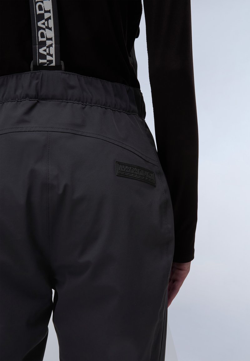Pantalones impermeables negros con tirantes ajustables, con un parche de logo texturizado en la parte trasera, tela suave y cinturilla elástica.
