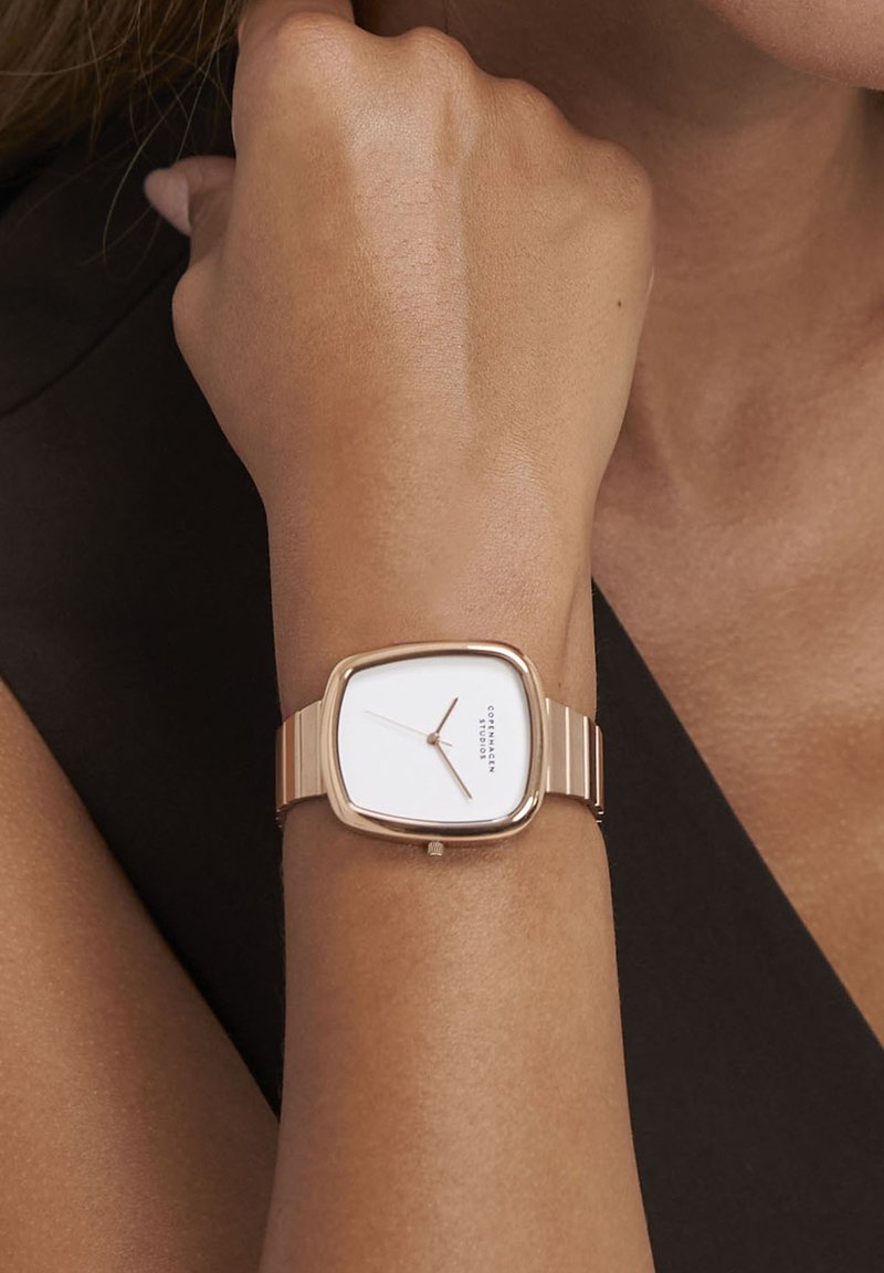 Orologio quadrato con quadrante bianco, cinturino in metallo dorato e profilo sottile. Presenta un design minimalista e un movimento a tre lancette.