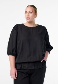 Melna blūze ar apaļu kakla izgriezumu, īsām puffed piedurknēm un savilktu ruffles apakšmalu. Gluda tekstūra ar vertikālu svītru rakstu.