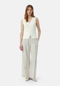 Witte mouwloze gebreide top met een V-hals en knoopsluiting, gecombineerd met lichtbeige wijde pantalon. Gladde textuur, minimaal ontwerp.