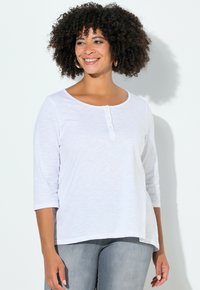 Haut blanc à manches longues avec un col rond, doté d'un demi-patine à boutons, fabriqué dans un tissu léger à texture lisse.