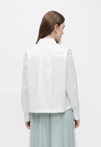 Valkoinen lyhyt takki kauluksella, napit takana ja pitkät hihat, yhdistetty vaaleansiniseen laskostettuun hameeseen.