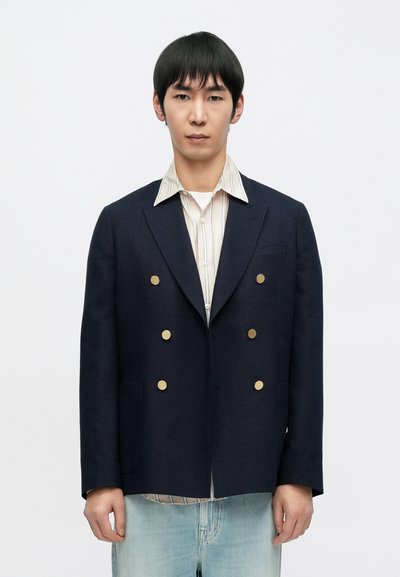 Marineblauer, doppelt geknöpfter Blazer mit goldenen Knöpfen, strukturiertem Stoff und Revers mit Kerben, getragen über einem gestreiften Hemd mit weißer Basis.