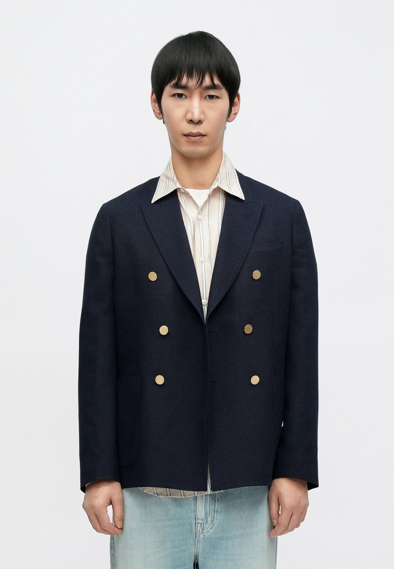 Granatowa dwurzędowa marynarka z złotymi guzikami, teksturowaną tkaniną i klapami z wycięciami, noszona na paskowanej koszuli z białą bazą.