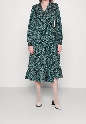 Femme portant une robe portefeuille verte à manches longues avec des taches blanches et un ourlet volanté, des chaussettes beiges et des chaussures crème à talon bloc carré.