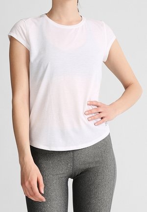 Kobieta ubrana w prosty biały, dopasowany t-shirt z krótkim rękawem oraz wysokiej talii szare legginsy, stojąca z jedną ręką na biodrze.