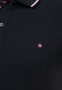 Polo negro con una tela texturizada, con un cuello a rayas rosas y un pequeño logotipo de estrella bordado en rosa en el pecho.