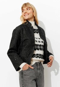 Veste courte noire à sequins avec un design structuré, portée sur un pull blanc avec un motif géométrique noir, associée à un jean gris.