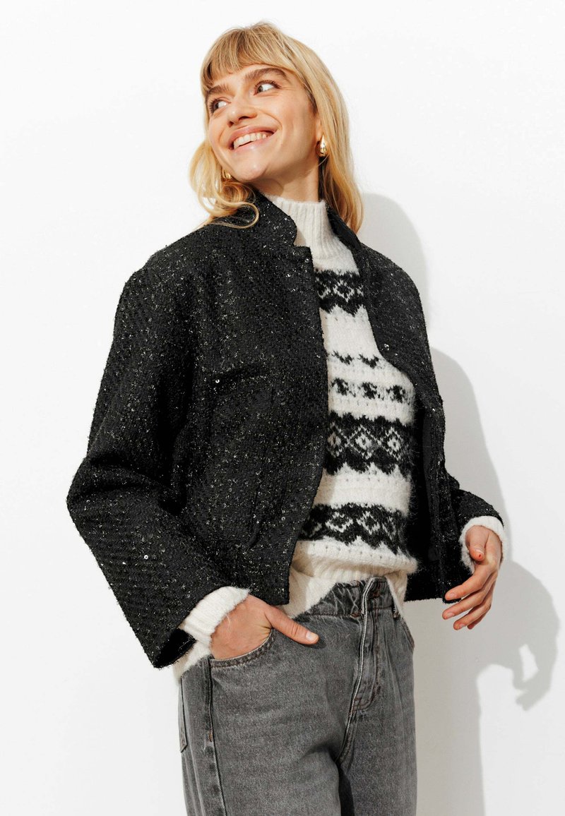 Veste courte noire à sequins avec un design structuré, portée sur un pull blanc avec un motif géométrique noir, associée à un jean gris.