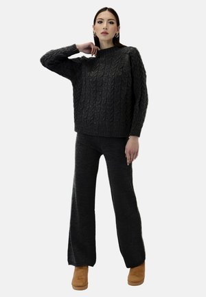 Donna che posa con maglione a trecce grigio scuro e pantaloni larghi in maglia, indossando stivaletti marrone chiaro su sfondo neutro.