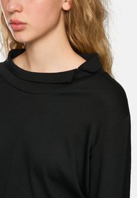 Schwarzes Langarmoberteil mit drapiertem Kragen und strukturiertem Stoff, mit lockerer Passform und sichtbaren Nähten entlang der Nähte.