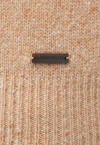 Suéter de punto beige con un patrón texturizado; presenta una etiqueta de tela negra rectangular con "DSQUARED2" grabado en el centro.