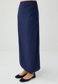 Marineblaue Maxi-Rock mit seitlichen Raffungselementen, aus einem glatten, leichten Stoff. Kombiniert mit schwarzen flachen Schuhen.
