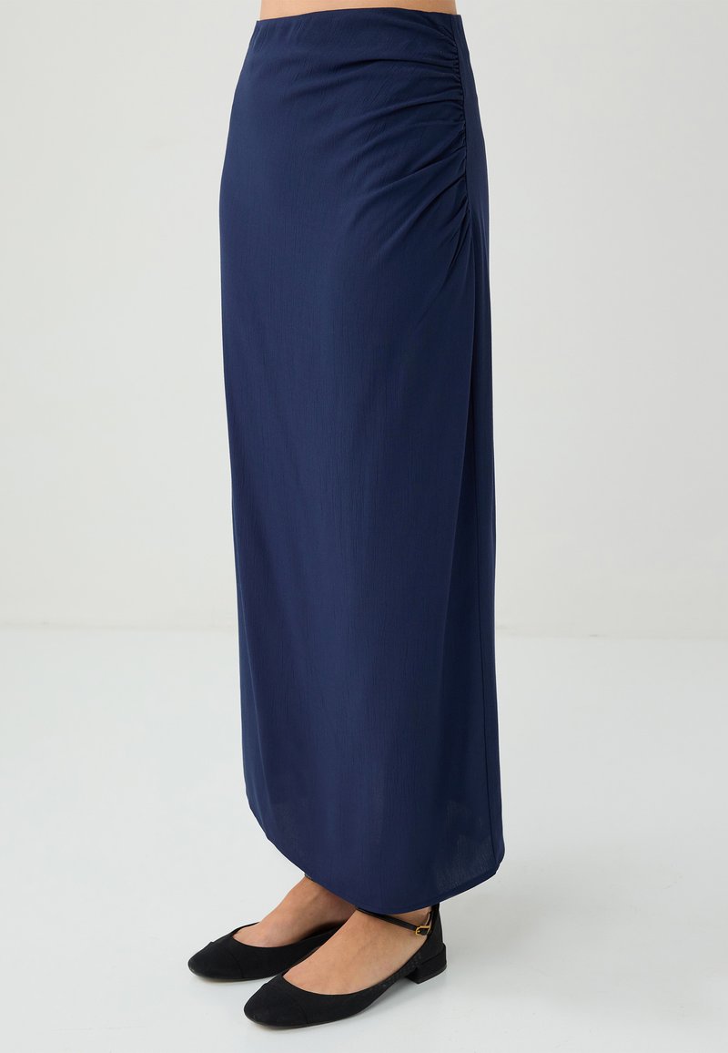 Marineblaue Maxi-Rock mit seitlichen Raffungselementen, aus einem glatten, leichten Stoff. Kombiniert mit schwarzen flachen Schuhen.