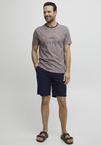 Short-sleeve, maroon heather t-shirt with orange graphic text, paired with navy shorts and brown sandals. Casual, relaxed fit.