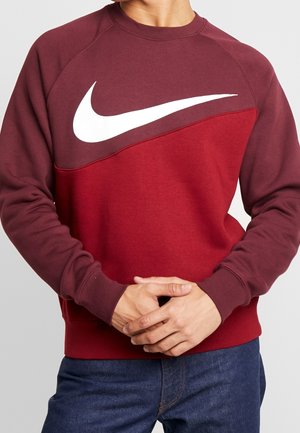 Osoba nosząca dwukolorową bordową i czerwoną bluzę z dużym białym logo Nike swoosh na piersi, z rękami splecionymi z przodu.