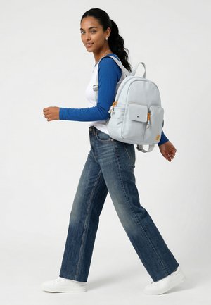 Kvinde går iført en blå og hvid langærmet skjorte, blå jeans, hvide sneakers og bærer en lysegrå rygsæk med forlommer.