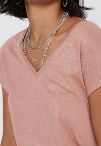 Top rosa de manga corta con diseño de escote en V. Hecho de una tela texturizada y brillante. Collares de cadena plateada en capas visibles como accesorios.