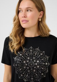 Sort t-shirt med et metalisk sølv cirkulært blomsterdesign, der indeholder prikker og sværmer. Rund hals og korte ærmer. Blødt stof.
