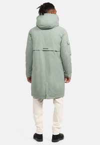 Parka lung, de culoare mint, cu glugă, având o ventilație pe spate, buzunare laterale și o etichetă cu logo, purtat cu pantaloni de culori deschise și cizme negre.
