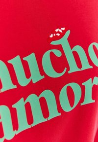 Czerwony bawełniany t-shirt z dużym zielonym napisem "Mucho" oraz wyszywaną czerwoną i białą papryczką chili nad napisem.