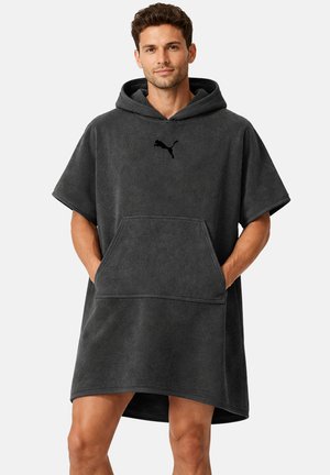 Mann trägt einen dunkelgrauen Kapuzenhandtuch-Poncho mit kurzen Ärmeln, Kängurutasche und Puma-Logo, steht mit den Händen in den Taschen.