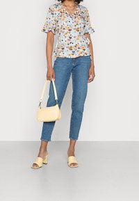 Blusa a stampa floreale con maniche corte, sfondo azzurro, abbinata a jeans blu aderenti. Borsa gialla in tessuto e tacchi aperti coordinati.