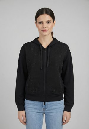 Giovane donna con capelli scuri raccolti in una coda di cavallo indossa una felpa nera con zip con piccoli borchie decorative e jeans azzurri chiari, in piedi in modo neutro.