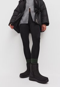 Botas negras aislantes con cierre de cremallera, tejido texturizado y suela gruesa. Combinadas con leggings negros y una blusa de punto gris.