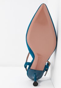 Chaussure à talon haut en daim bleu à bout pointu vue de la semelle, reposant sur son talon contre une surface et un mur blancs.