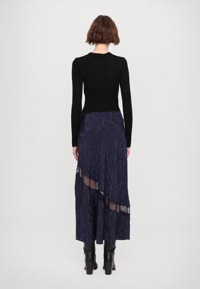 Schwarzes, geripptes Langarmoberteil kombiniert mit einem langen, marineblauen Rock mit Spitzenakzenten und einem strukturierten Muster, ergänzt durch kniehohe schwarze Stiefel.
