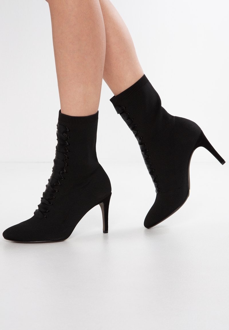 Bottes noires à talons hauts, chevilles, à bout pointu avec un détail de ruban lacé, portées sur des jambes nues sur fond blanc.