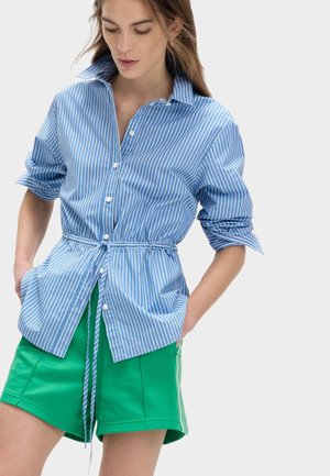 Vrouw die een blauw-wit gestreept overhemd met knopen draagt, met een trekkoord in de taille en groene korte broek, handen in de zakken, naar beneden kijkend.