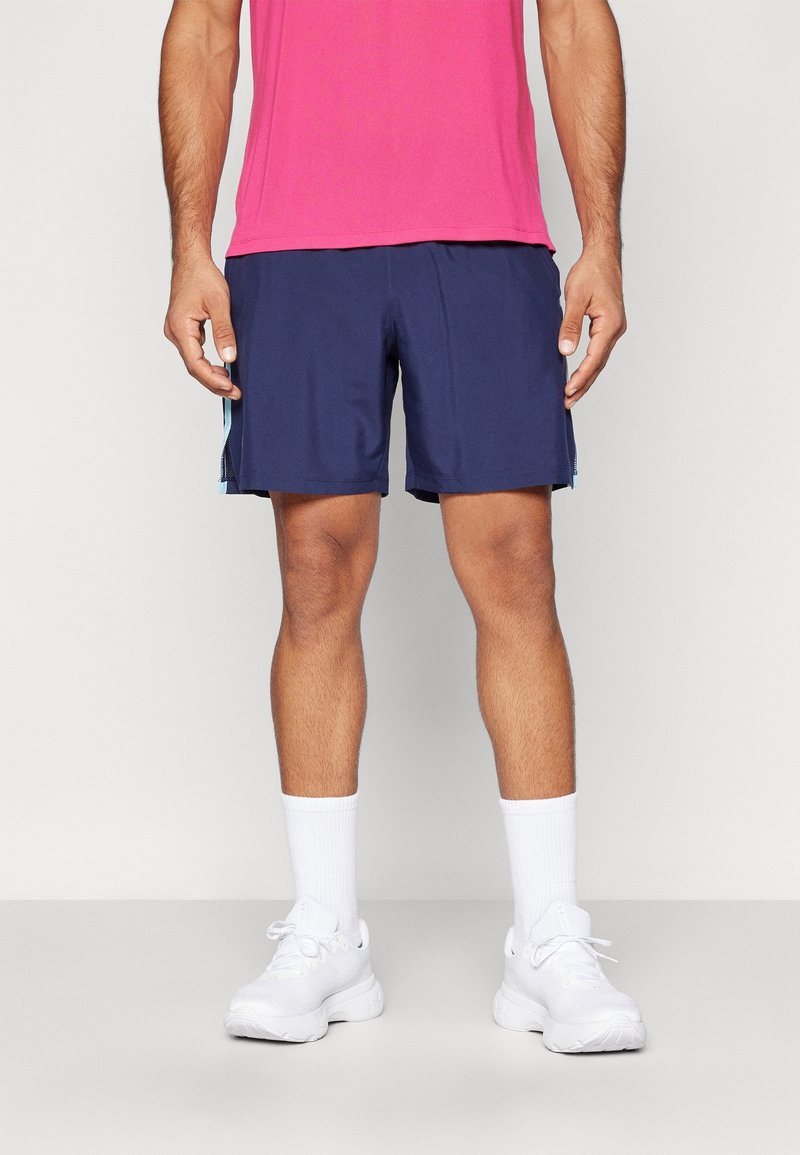 Man som har på sig marinblå träningstights, vita ankelsockor och vita sneakers, stående mot en neutral bakgrund.