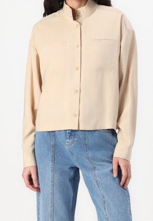 Chemise beige à manches longues boutonnée avec col montant portée avec un jean en denim bleu taille haute avec coutures visibles à l'avant.