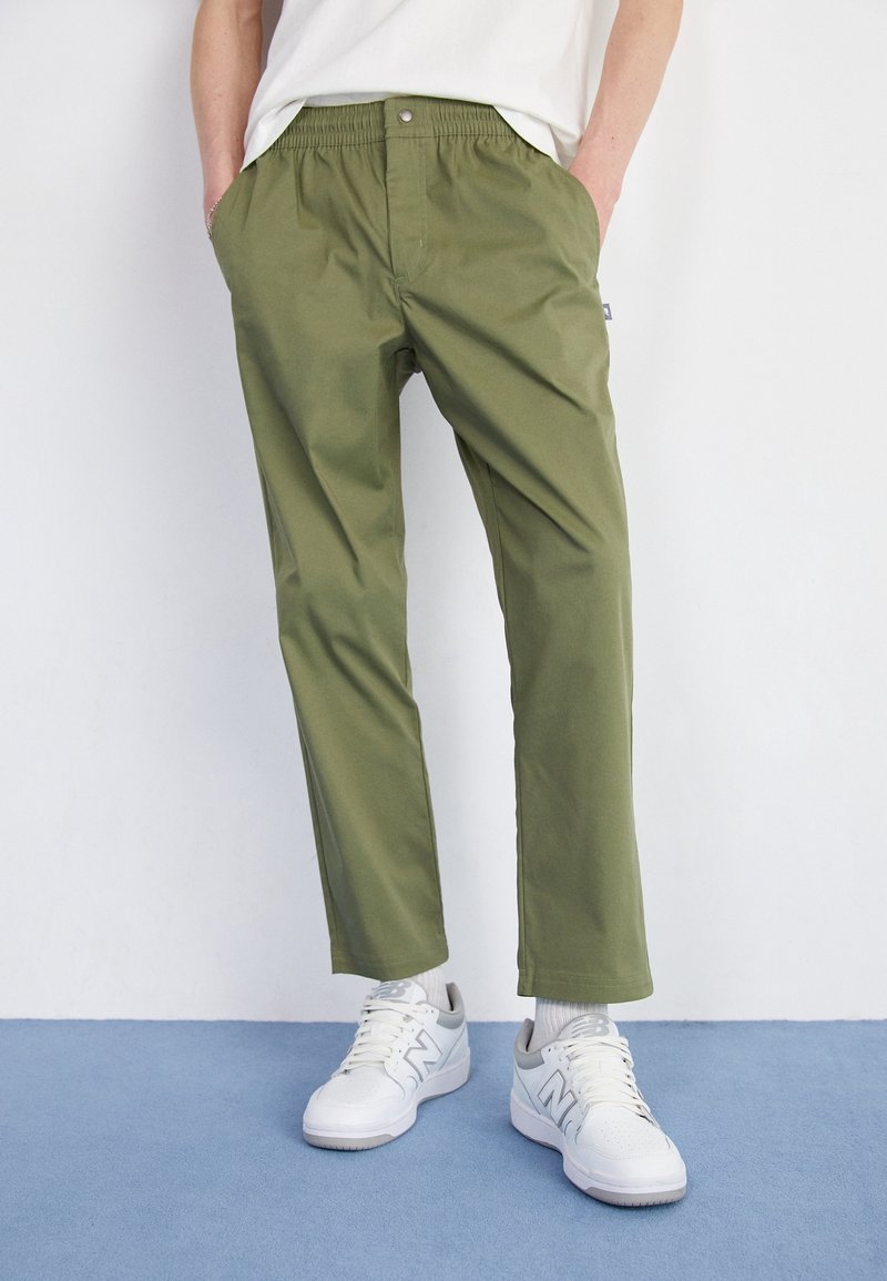 Personne portant un pantalon vert olive à taille élastique, des baskets blanches et des chaussettes blanches, debout sur un sol bleu clair contre un mur blanc.