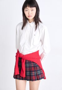 Camisa blanca de botón con un pequeño logo, suéter de punto rojo atado a la cintura y falda de cuadros plisada en tonos azul oscuro, rojo y verde.